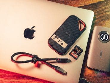 A silver Apple MacBook resting on a wooden desk. Placed on top of and beside the laptop are a SanDisk 2TB portable SSD labeled "MUDI 1", a 128GB SanDisk SD card, a short braided USB-C cable, and a gray llano-brand external hub or card reader