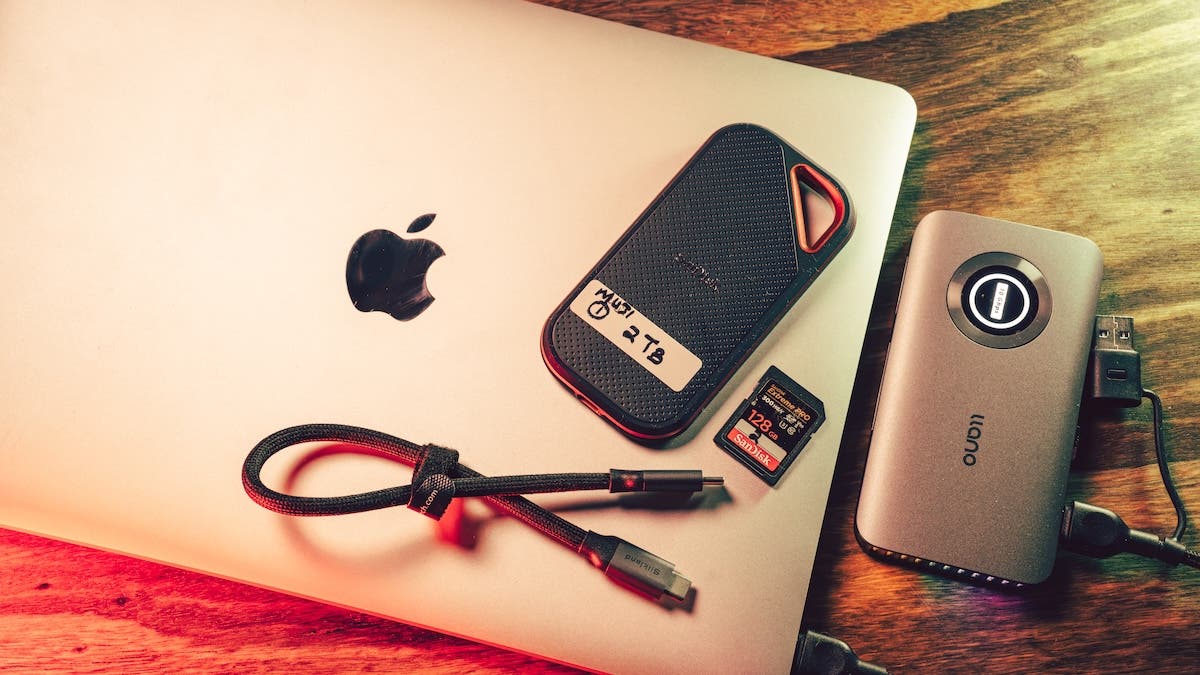 A silver Apple MacBook resting on a wooden desk. Placed on top of and beside the laptop are a SanDisk 2TB portable SSD labeled "MUDI 1", a 128GB SanDisk SD card, a short braided USB-C cable, and a gray llano-brand external hub or card reader