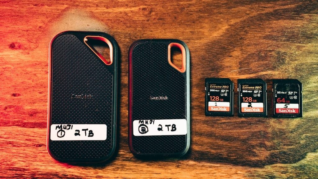 Two SanDisk portable SSDs with white handwritten labels reading "MUDI 1 2TB" and "MUDI 2 2TB", arranged next to three SanDisk SD cards (two 128GB Extreme PRO and one 64GB Extreme) on a wooden surface