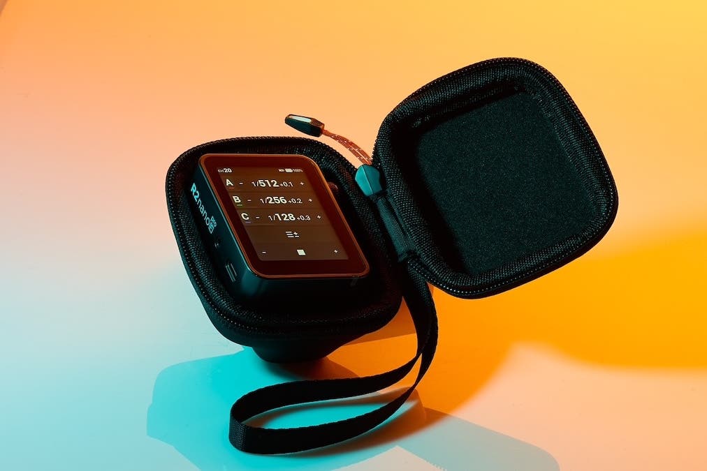 A final close-up product photograph of a small, black Flashpoint wireless flash trigger resting inside its open, semi-hard zippered carrying case. The object is dramatically lit with contrasting, vibrant gradients of orange and cyan.
