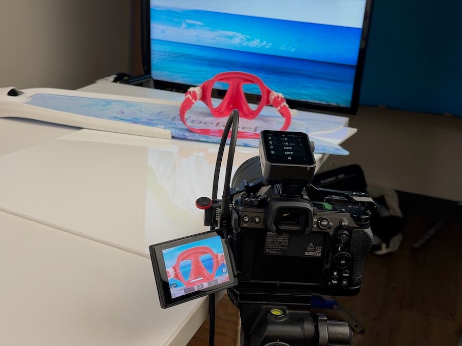 A behind-the-scenes look at a product photo shoot for a pink snorkeling mask. A camera on a tripod is aimed at the mask, which rests in front of a computer monitor displaying a tropical beach scene. The camera's articulated LCD screen shows the final framed shot, using the monitor as a realistic background.
