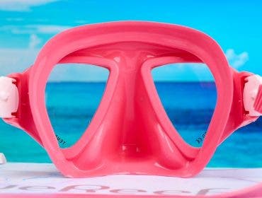 A final close-up product photograph of a bright pink snorkeling mask. Similar to the purple mask, a bright blue ocean and sky scene is perfectly framed through the clear lenses, creating the illusion that it is resting on a beach.