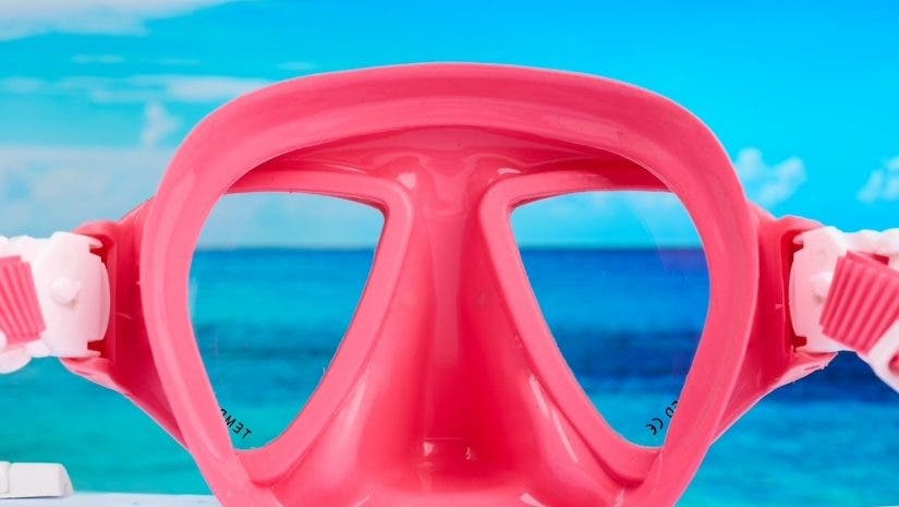 A final close-up product photograph of a bright pink snorkeling mask. Similar to the purple mask, a bright blue ocean and sky scene is perfectly framed through the clear lenses, creating the illusion that it is resting on a beach.