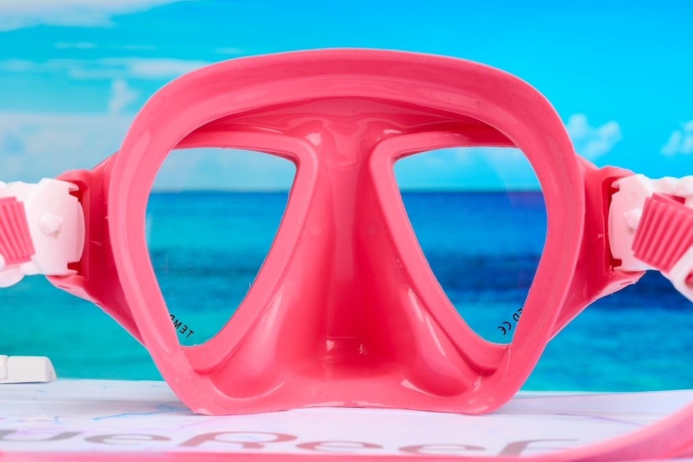 A final close-up product photograph of a bright pink snorkeling mask. Similar to the purple mask, a bright blue ocean and sky scene is perfectly framed through the clear lenses, creating the illusion that it is resting on a beach.