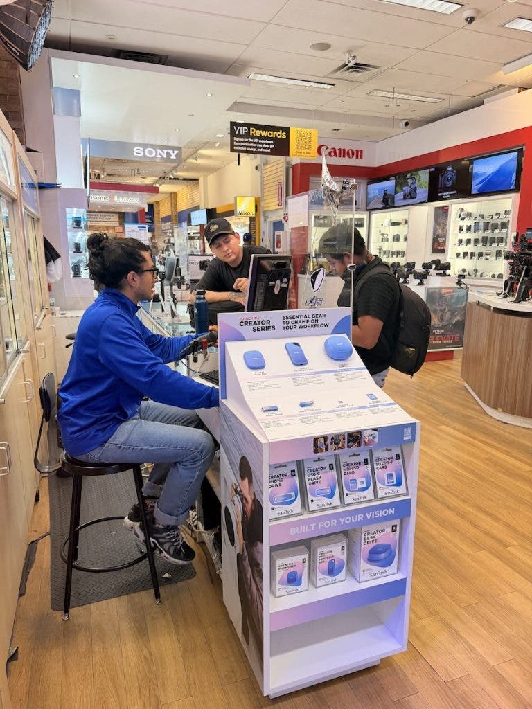 A customer in a blue shirt sits at a counter in a brightly lit electronics store, conversing with two employees behind a clear plastic barrier. In the foreground is a retail display for SanDisk Creator Series products, and overhead signs for Sony, Canon, and Panasonic are visible in the background