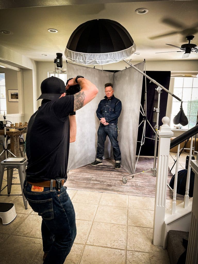 Behind-the-scenes view of photographer Ian Spanier taking a portrait of actor Robert Patrick, who stands in head-to-toe denim in front of a studio backdrop inside a home set up with professional studio lighting and a large softbox umbrella
