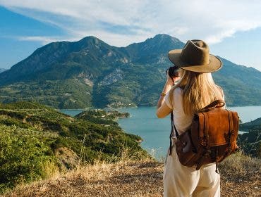 A woman with a hat and leather backpack taking a photo of a scenic lake surrounded by green mountains on a sunny day.