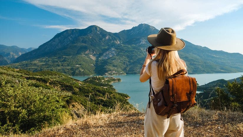 A woman with a hat and leather backpack taking a photo of a scenic lake surrounded by green mountains on a sunny day.