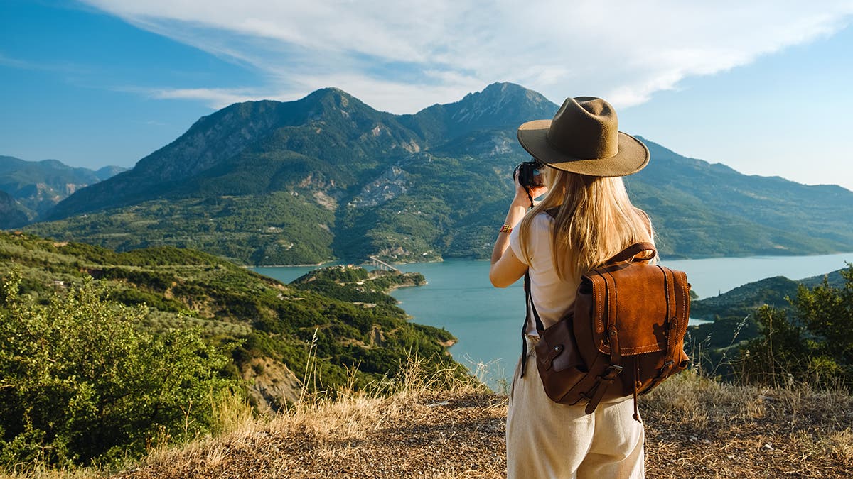 A woman with a hat and leather backpack taking a photo of a scenic lake surrounded by green mountains on a sunny day.