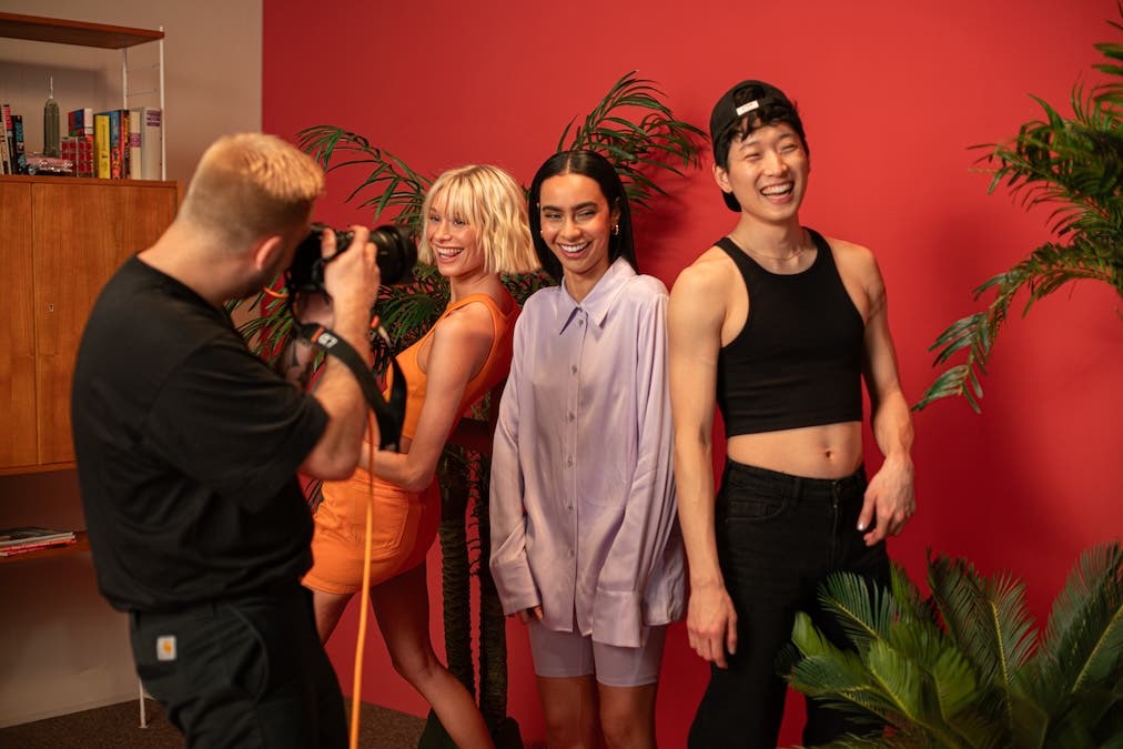 Photographer shooting three smiling models standing closely together against a vibrant red backdrop flanked by tropical indoor potted plants