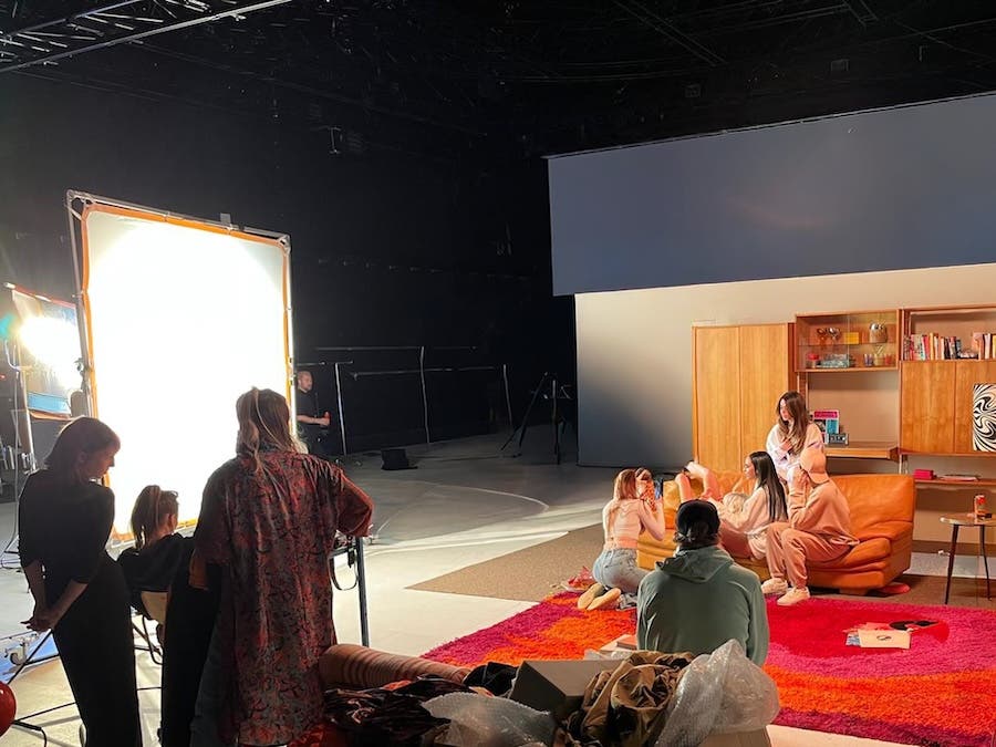 Behind-the-scenes view of a film set showing a production crew surrounding a large 12x12 diffused lighting frame pointing toward models sitting on a couch in a retro living room set