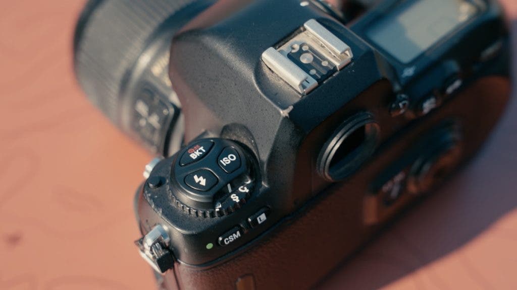 A close-up shot of the top left panel of a black Nikon film camera, highlighting the hot shoe mount and the control dial featuring ISO, flash, and bracketing buttons