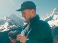A man wearing a black cap and blue fleece jacket stands outdoors in front of snow-capped mountains, looking down at the black Nikon camera he is holding