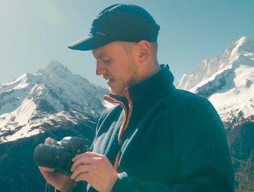 A man wearing a black cap and blue fleece jacket stands outdoors in front of snow-capped mountains, looking down at the black Nikon camera he is holding