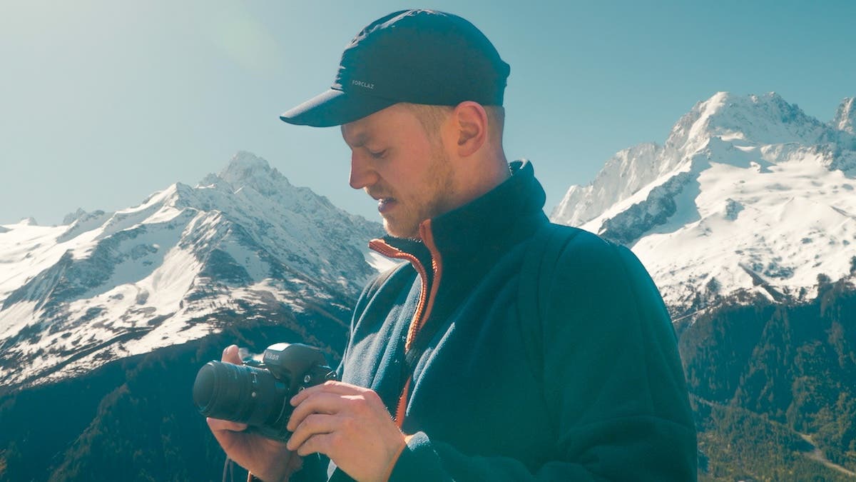 A man wearing a black cap and blue fleece jacket stands outdoors in front of snow-capped mountains, looking down at the black Nikon camera he is holding