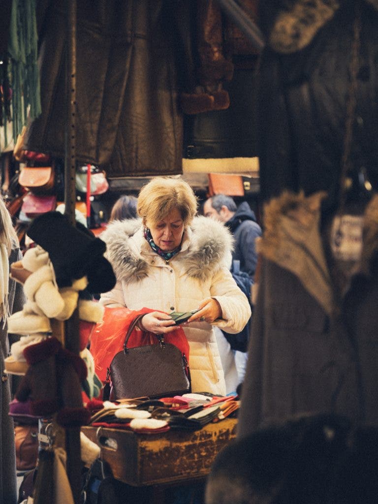 A woman in a white winter coat browsing small leather goods at an outdoor market stall, candidly captured by invisible street photographer Muji using a 67mm mid-range zoom lens.