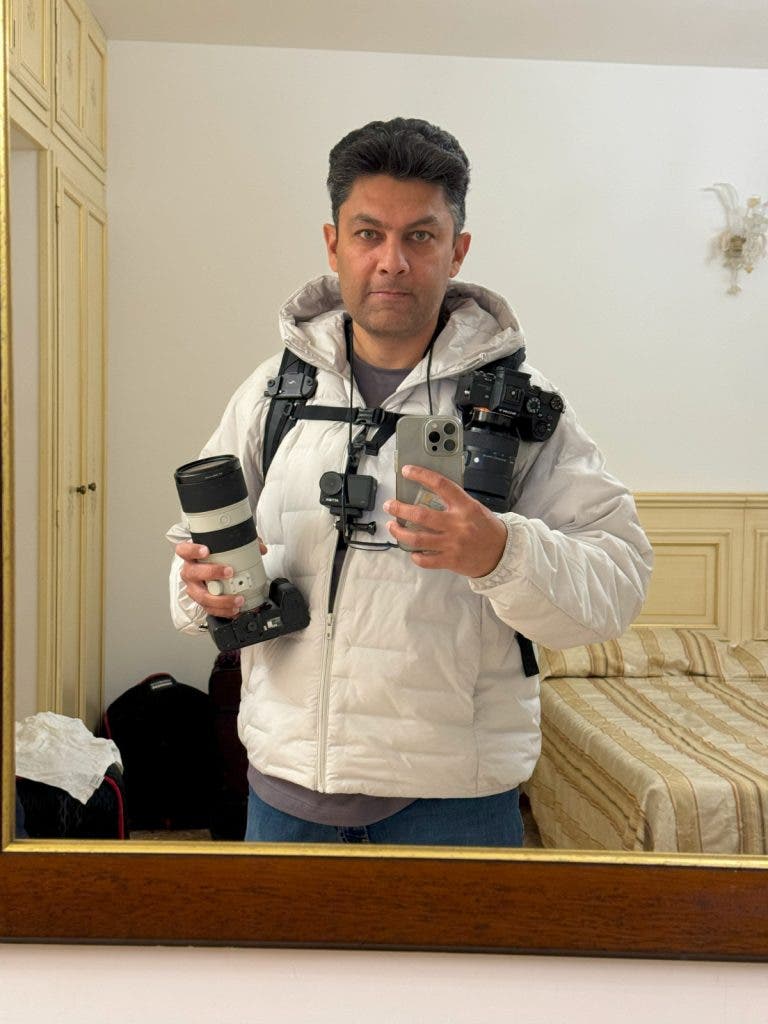 Mirror selfie of invisible street photographer Muji wearing a white puffer jacket strapped with multiple professional cameras, telephoto lenses, and a smartphone rigged for photography.