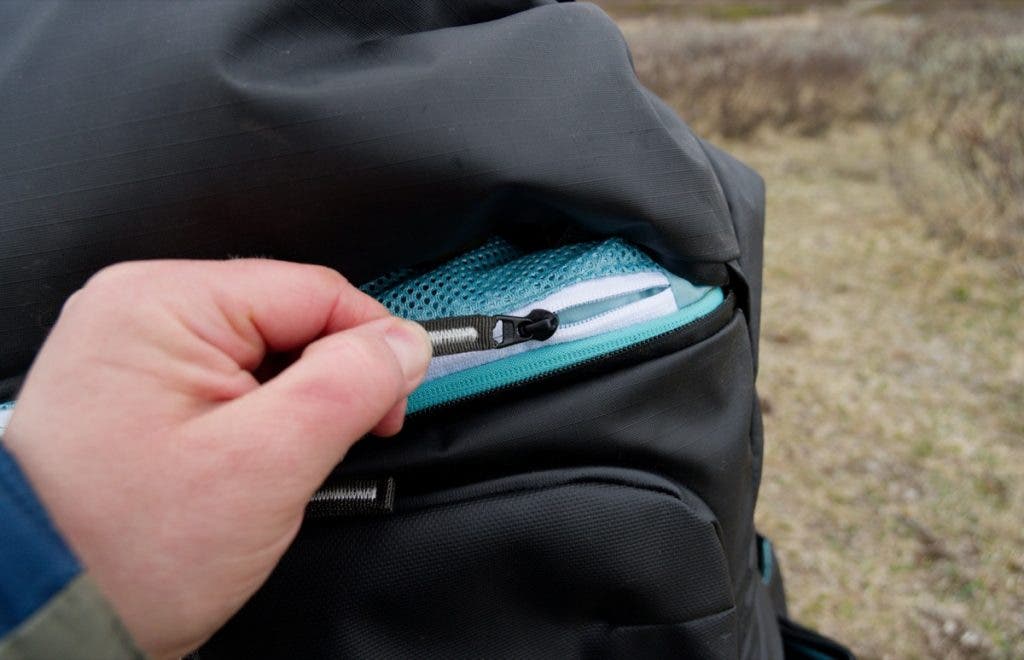 A close-up shot of a hand opening a zipper on a black Shimoda Action X70 HD backpack to reveal a light blue mesh pocket inside.