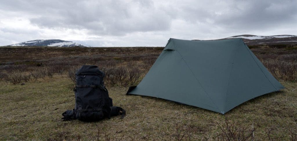 A black Shimoda Action X70 HD 70L backpack sitting on grassy ground next to a pitched grey camping tent in an open mountain valley.