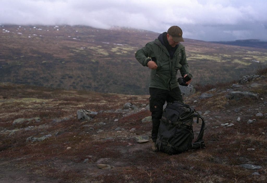 Peter Dam is standing outdoors in mountain terrain, holding a camera next to a black Shimoda Action X70 HD 70L backpack resting on the ground.