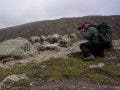 Photographer wearing a large black backpack crouched in a rocky mountain landscape taking a picture with a camera.