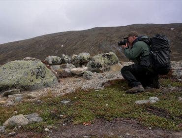 Photographer wearing a large black backpack crouched in a rocky mountain landscape taking a picture with a camera.