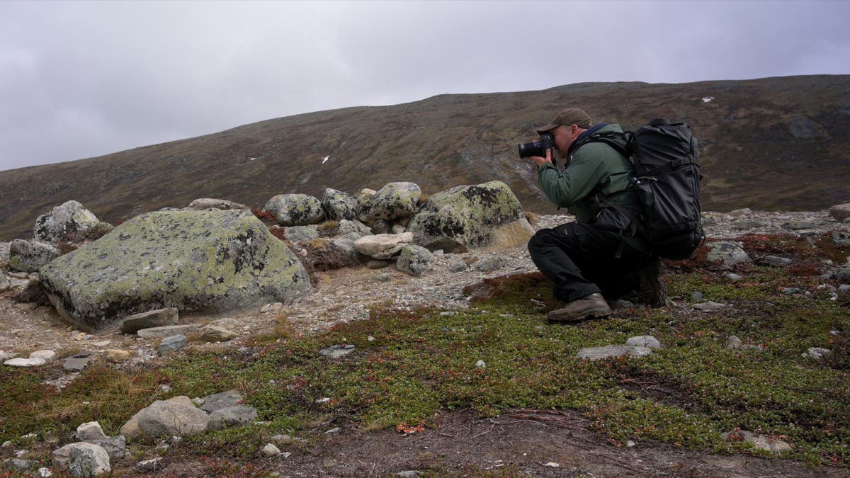 Photographer wearing a large black backpack crouched in a rocky mountain landscape taking a picture with a camera.