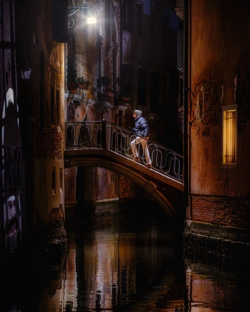 A hooded pedestrian crossing a stone bridge over a Venetian canal illuminated by streetlights at night, shot from a dark alley by invisible street photographer Muji.