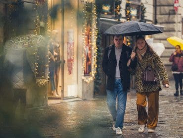 A couple sharing a black umbrella on a rainy, festive city street lined with warm shop lights, captured candidly through foreground foliage by invisible street photographer Muji.