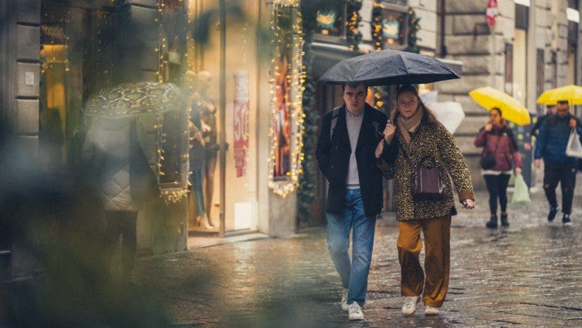 A couple sharing a black umbrella on a rainy, festive city street lined with warm shop lights, captured candidly through foreground foliage by invisible street photographer Muji.
