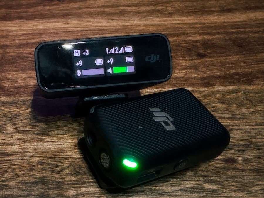 A black DJI wireless microphone transmitter and receiver resting on a wooden surface. The receiver's digital screen displays audio levels, signal strength, and battery icons, while the transmitter unit in the foreground features a glowing green indicator light and the DJI logo.