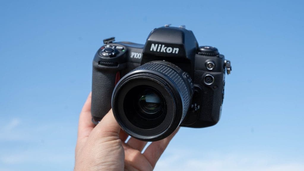 A hand holds up a black Nikon F100 35mm film camera with an attached lens against a clear blue sky