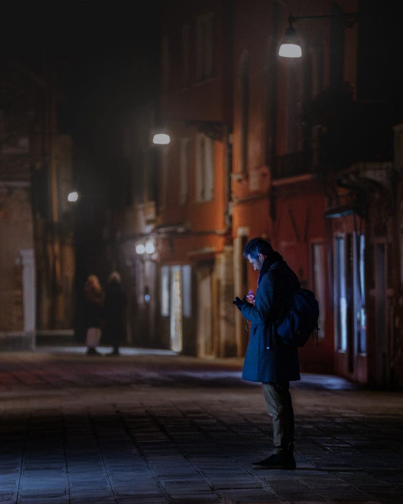 A candid nighttime shot by invisible street photographer Muji of a man standing on a dimly lit European city street while looking down at his phone, taken with a 178mm zoom lens.