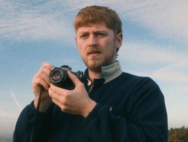 A bearded man holding a Canon film camera outdoors, looking intently toward the camera against a bright, partly cloudy sky