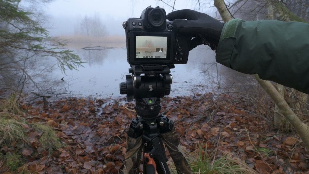 A close-up of a gloved hand operating a black camera mounted on a camouflage-patterned tripod, set up on a shore with brown autumn leaves overlooking a foggy lake
