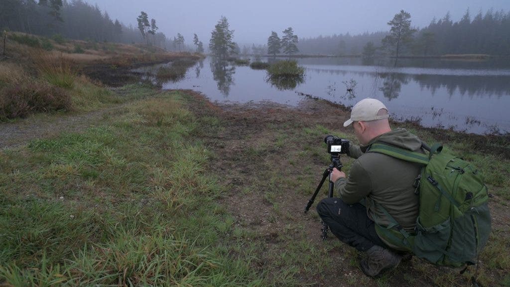 A photographer wearing a green jacket and backpack kneels in a grassy area, adjusting a camera on a tripod pointed toward a fog-covered lake bordered by pine trees