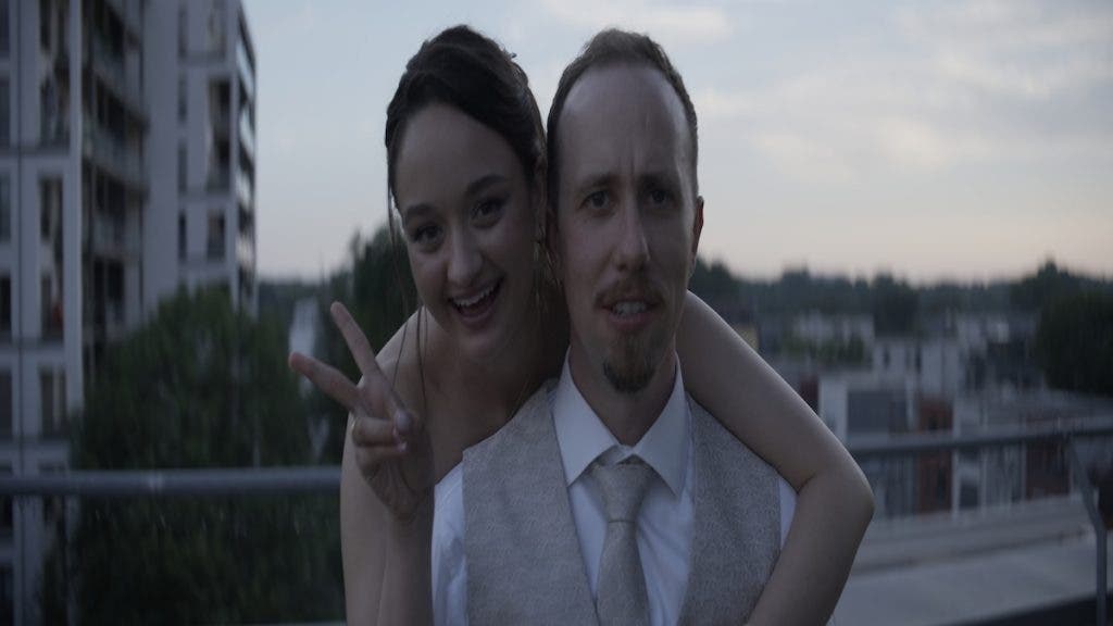 A woman in a white dress makes a peace sign while embracing a man from behind, set against a dusky city backdrop