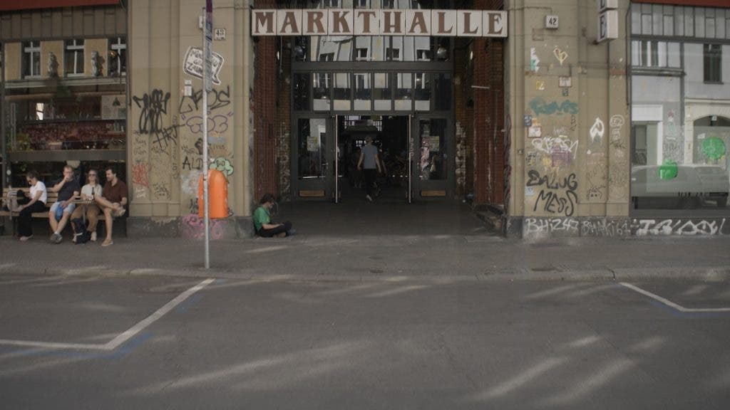 A wide-angle view of a building entrance labeled "MARKTHALLE," showing graffiti-covered brick pillars, patrons at a cafe to the left, and a person sitting on the ground