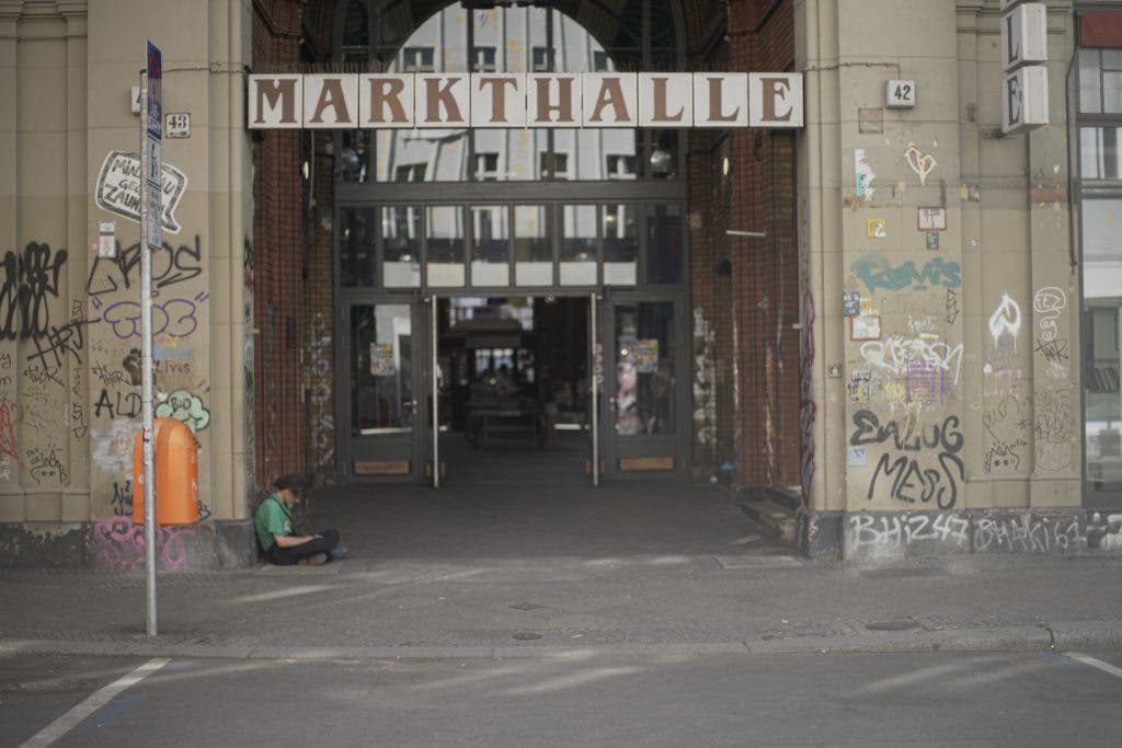A standard-aspect view of the "MARKTHALLE" building entrance, focusing on the doors, the graffiti on the right pillar, and a person sitting on the ground