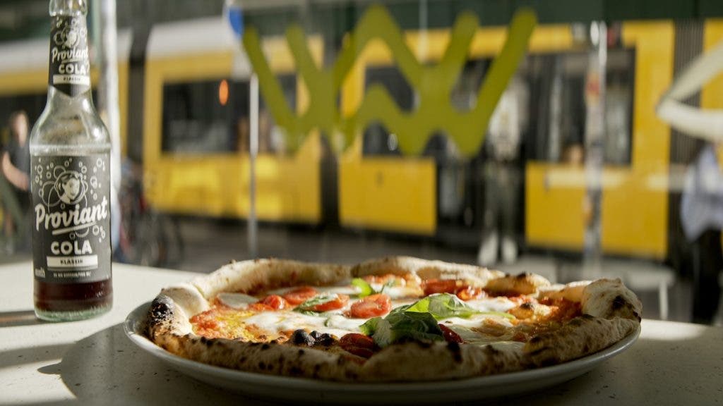 A bottle of Proviant Cola and a Margherita pizza on a counter, with a blurred yellow train passing by in the background window
