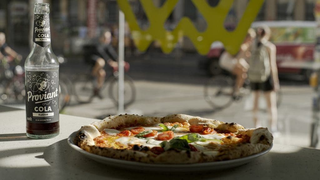 A bottle of Proviant Cola and a Margherita pizza on a counter, with blurred pedestrians and cyclists moving past the window in the background