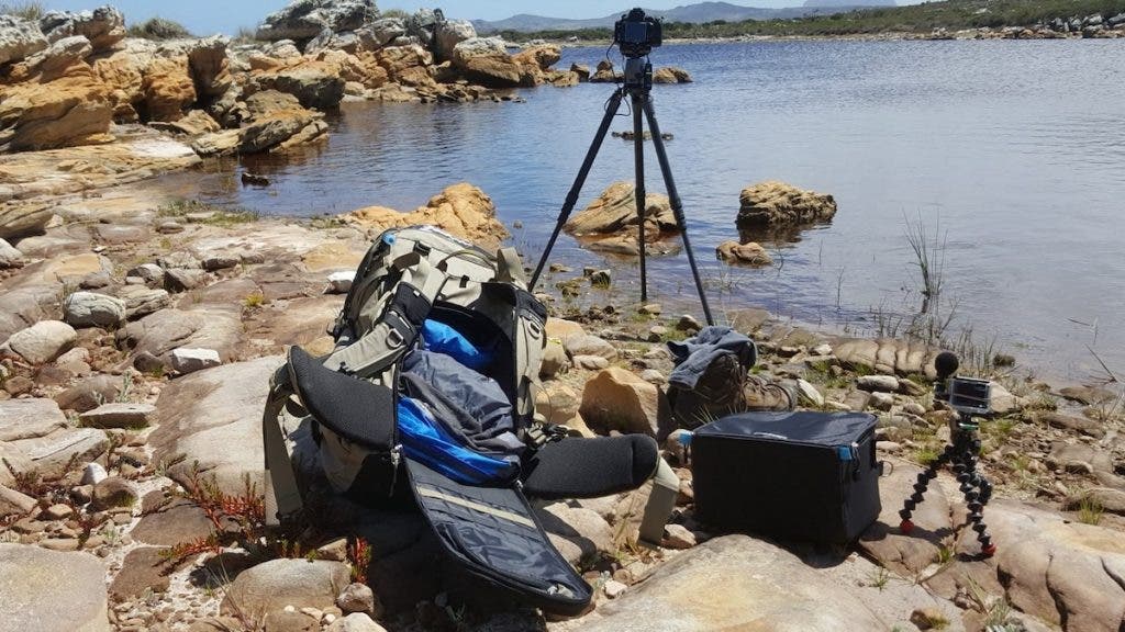 A behind-the-scenes camera setup on a rocky shoreline next to the water, featuring an open backpack, a primary camera on a tall tripod, and a GoPro action camera mounted on a small, flexible tripod