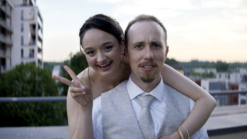 A smiling woman holds up a peace sign while hugging a man from behind, both looking directly at the camera outdoors at dusk