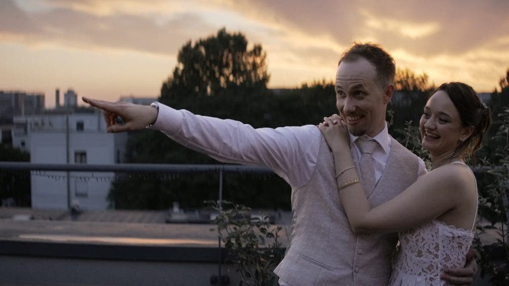 A man in a beige vest points off-camera to the left while a woman in a white lace dress hugs him from behind, backlit by a sunset