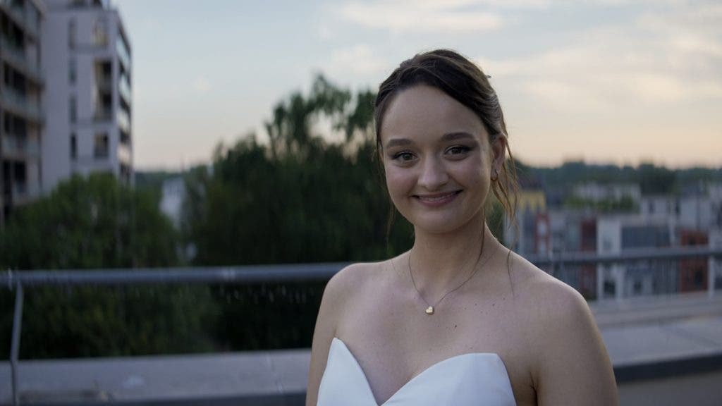 A portrait of a smiling woman wearing a white sweetheart-neckline dress and a small heart necklace, standing on a rooftop or balcony at dusk
