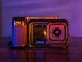 A close-up of a GoPro action camera mounted inside a black protective cage with a side handle grip, set against a vibrantly lit purple and orange background