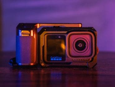 A close-up of a GoPro action camera mounted inside a black protective cage with a side handle grip, set against a vibrantly lit purple and orange background
