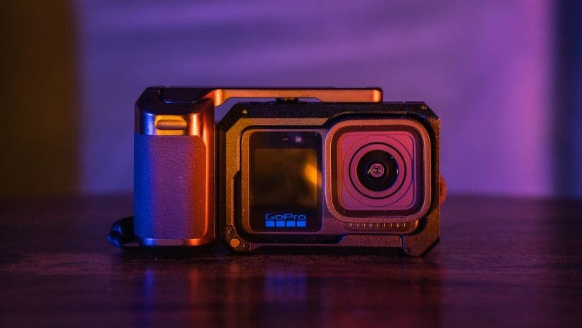 A close-up of a GoPro action camera mounted inside a black protective cage with a side handle grip, set against a vibrantly lit purple and orange background