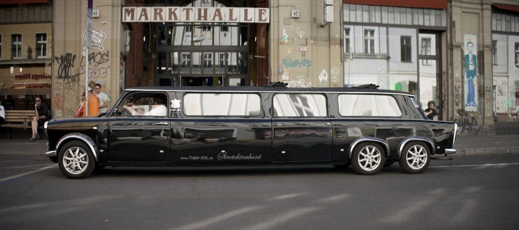 A black multi-axle stretch limousine modified to look like a classic Trabant parked in front of a graffiti-covered building entrance labeled "MARKTHALLE"