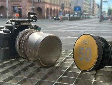 Two camera lenses resting on a metal grate with a blurred city intersection behind them; the left is a silver DZOFILM lens attached to a black camera cage, and the right is a black lens with a yellow Contax Zeiss 50 cap